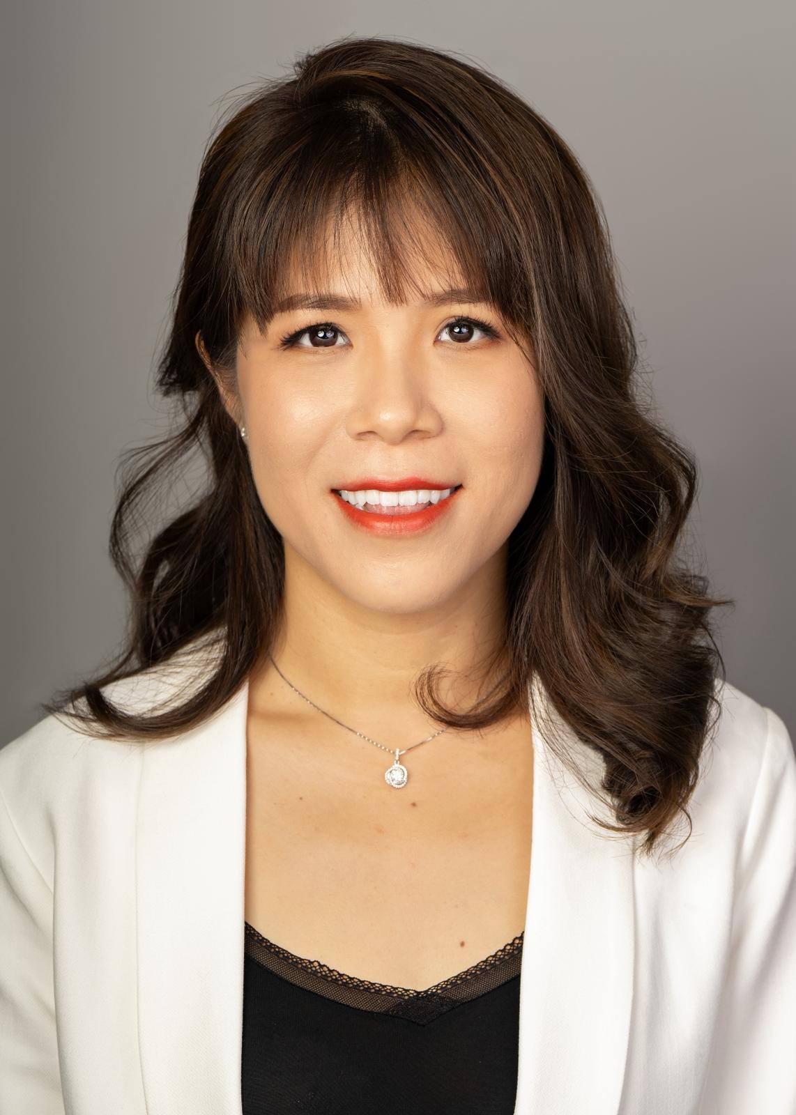 A woman wearing a white jacket over a black top, with wavy hair and a necklace, posing against a neutral background.