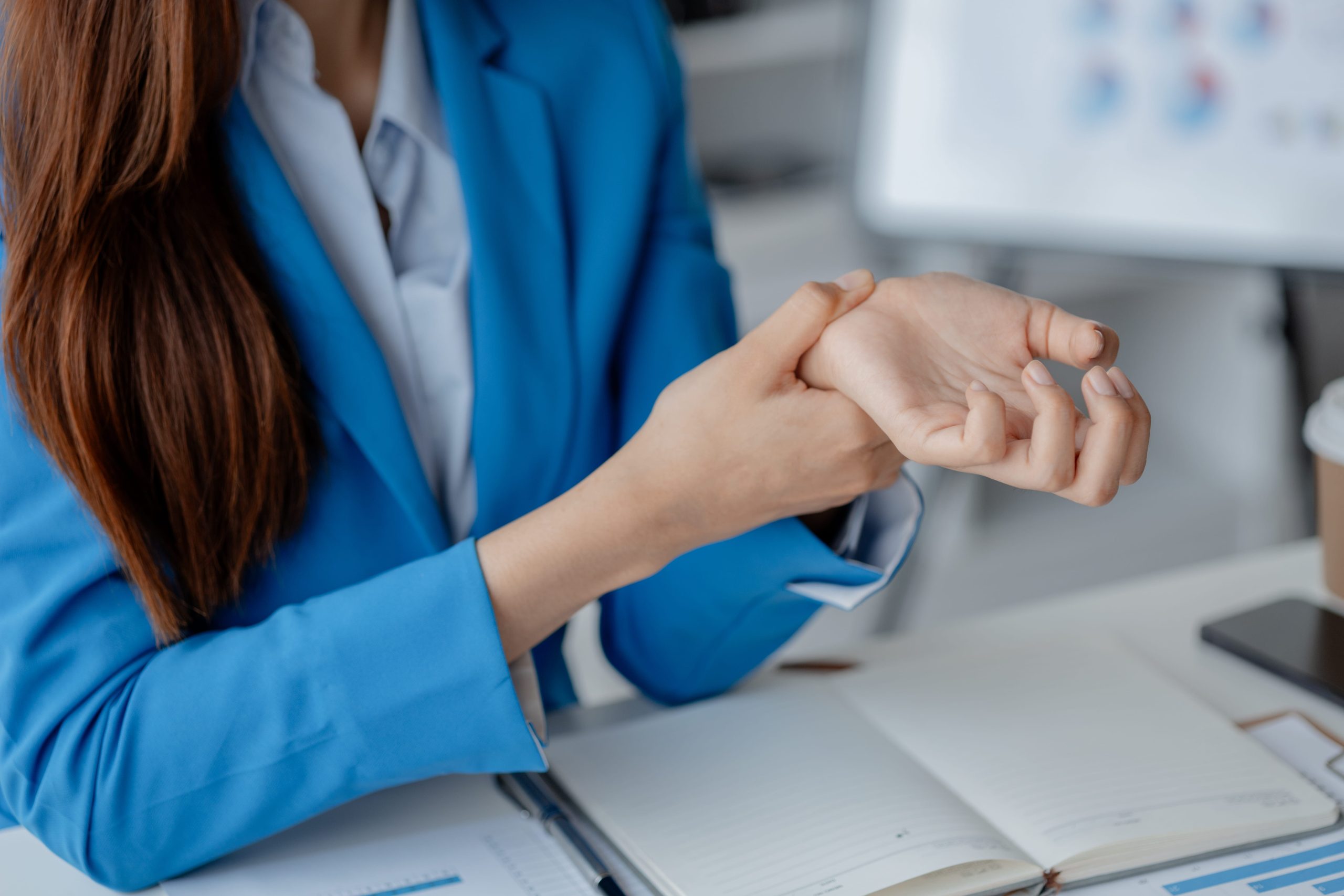 A woman wearing a blue blazer examines her wrist while seated at a desk filled with notes and a coffee cup.